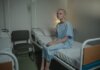 Либералы провалили законопроект о запрете MAiD при психических расстройствах A young woman in a hospital gown sits on a bed in a medical setting, conveying a somber mood.