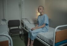 A young woman in a hospital gown sits on a bed in a medical setting, conveying a somber mood.