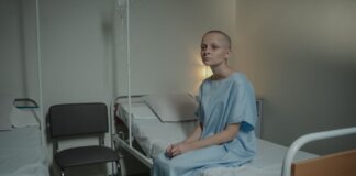 A young woman in a hospital gown sits on a bed in a medical setting, conveying a somber mood.