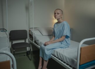 A young woman in a hospital gown sits on a bed in a medical setting, conveying a somber mood.