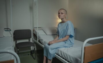 Либералы провалили законопроект о запрете MAiD при психических расстройствах A young woman in a hospital gown sits on a bed in a medical setting, conveying a somber mood.