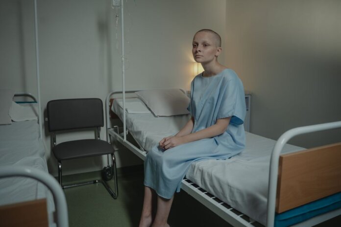 A young woman in a hospital gown sits on a bed in a medical setting, conveying a somber mood. A young woman in a hospital gown sits on a bed in a medical setting, conveying a somber mood.