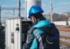 An engineer in a blue hard hat examines a fuse box on a railway track outdoors.