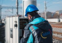 An engineer in a blue hard hat examines a fuse box on a railway track outdoors.
