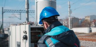 An engineer in a blue hard hat examines a fuse box on a railway track outdoors.