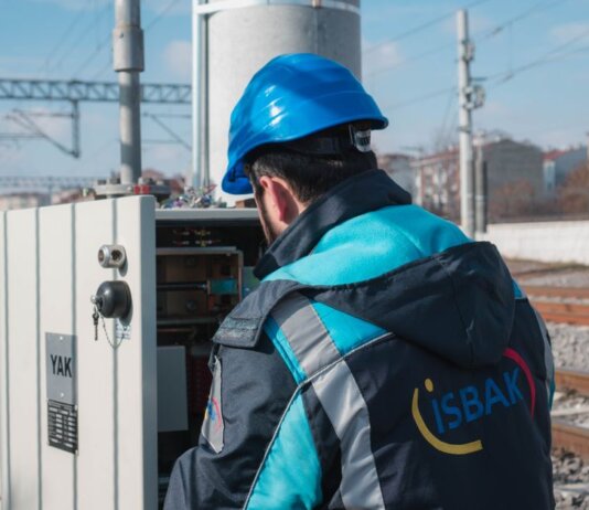 An engineer in a blue hard hat examines a fuse box on a railway track outdoors.