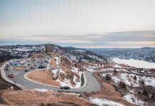 Automobiles riding on curvy asphalt roadway near grassy shore covered with snow against river and cloudy sky on winter day