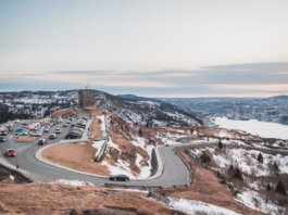 Automobiles riding on curvy asphalt roadway near grassy shore covered with snow against river and cloudy sky on winter day