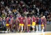 Basketball team huddles on court during a game timeout with audience in background.