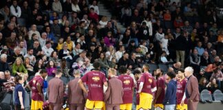 Basketball team huddles on court during a game timeout with audience in background.