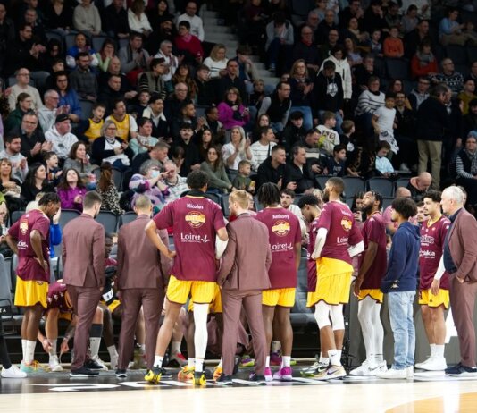 Basketball team huddles on court during a game timeout with audience in background.