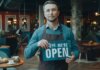 Cafe owner standing inside, holding a 'We're Open' sign, welcoming customers.