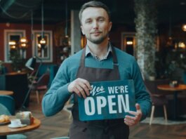 Cafe owner standing inside, holding a 'We're Open' sign, welcoming customers.