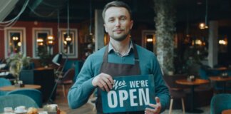 Cafe owner standing inside, holding a 'We're Open' sign, welcoming customers.