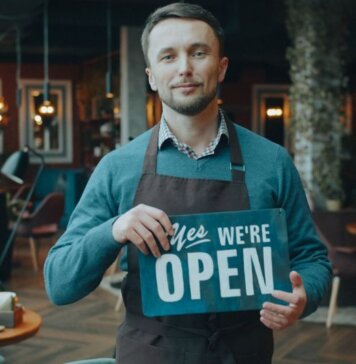 Cafe owner standing inside, holding a 'We're Open' sign, welcoming customers.
