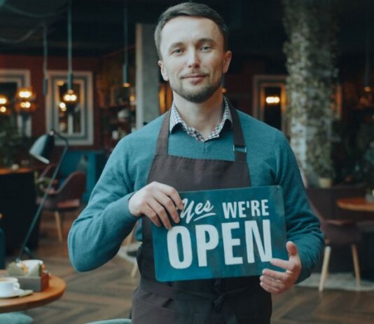 Cafe owner standing inside, holding a 'We're Open' sign, welcoming customers.
