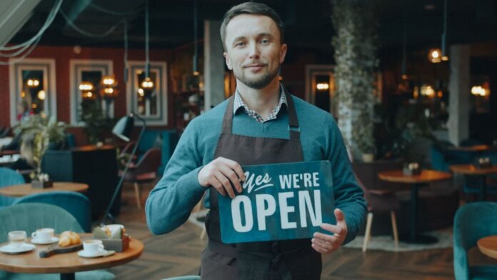 Cafe owner standing inside, holding a 'We're Open' sign, welcoming customers. Cafe owner standing inside, holding a 'We're Open' sign, welcoming customers.