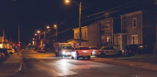 Cars with glowing lights parked on asphalt road near police car placed near residential building in small town in evening