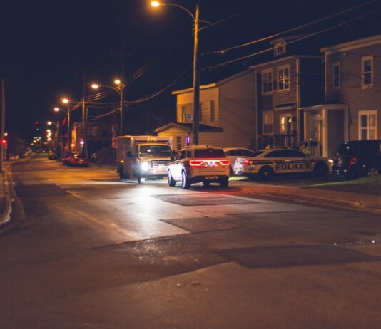 Cars with glowing lights parked on asphalt road near police car placed near residential building in small town in evening