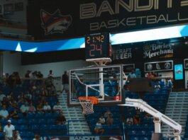 Close-up of a basketball hoop inside a stadium with a scoreboard and audience in the background.