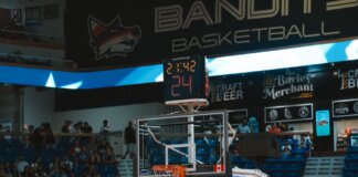 Close-up of a basketball hoop inside a stadium with a scoreboard and audience in the background.