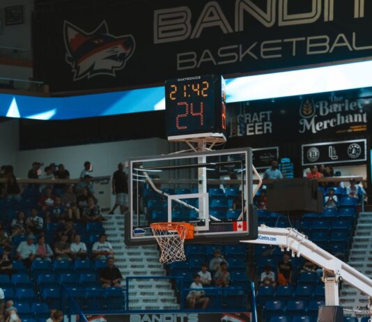 Close-up of a basketball hoop inside a stadium with a scoreboard and audience in the background.