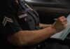 В Калгари двоим предъявлены обвинения в мошенничестве на $1 млн Close-up of a police officer writing in a notebook while sitting in a car, focused detail.