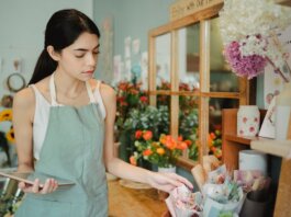 Concentrated female florist with dark hair standing in floral shop with tablet near flowers in wrapper while checking order during work