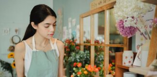 Concentrated female florist with dark hair standing in floral shop with tablet near flowers in wrapper while checking order during work