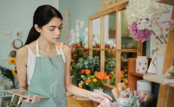 Concentrated female florist with dark hair standing in floral shop with tablet near flowers in wrapper while checking order during work