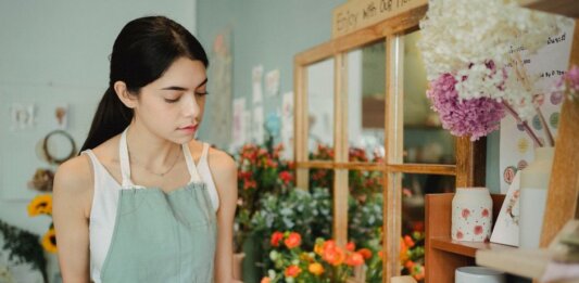 Concentrated female florist with dark hair standing in floral shop with tablet near flowers in wrapper while checking order during work