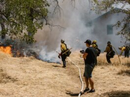Firefighters in protective gear combating a spreading wildfire near a home.
