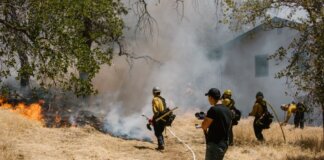 Firefighters in protective gear combating a spreading wildfire near a home.