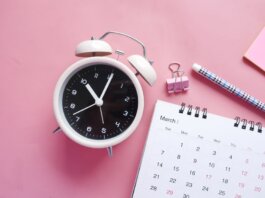 Flat lay of a pink-themed workspace featuring an alarm clock, calendar, pen, and paper clip on a pastel surface.