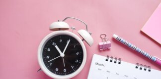 Flat lay of a pink-themed workspace featuring an alarm clock, calendar, pen, and paper clip on a pastel surface.