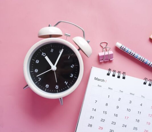 Flat lay of a pink-themed workspace featuring an alarm clock, calendar, pen, and paper clip on a pastel surface.