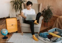 Man sitting on armchair planning travel, using laptop, with suitcases and globe nearby.