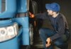 Mechanic in uniform checking a blue truck's wheel inside a garage workshop.