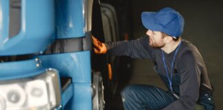 Mechanic in uniform checking a blue truck's wheel inside a garage workshop.