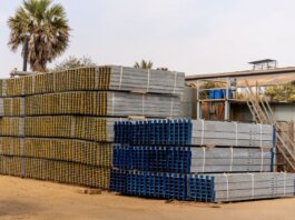 Piles of stacked metal beams in an industrial outdoor yard under a clear sky.