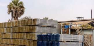 Piles of stacked metal beams in an industrial outdoor yard under a clear sky.