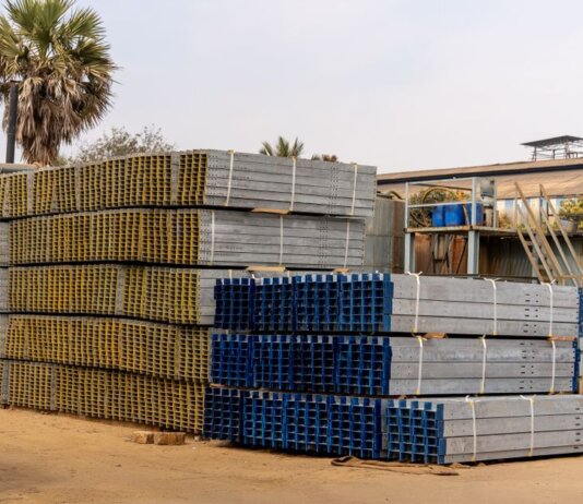 Piles of stacked metal beams in an industrial outdoor yard under a clear sky.