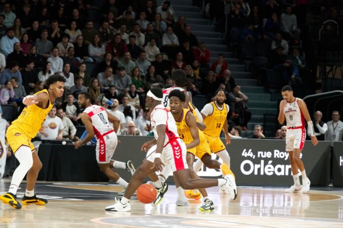 Players in action during an indoor basketball game, with a crowd watching intently. Players in action during an indoor basketball game, with a crowd watching intently.