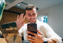 WhatsApp открыл звонки для всех: присоединиться теперь можно без аккаунта Smiling young man making a video call on his smartphone while sitting with a laptop in a modern office.