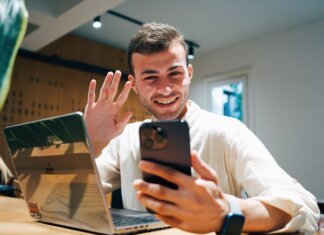 Smiling young man making a video call on his smartphone while sitting with a laptop in a modern office.
