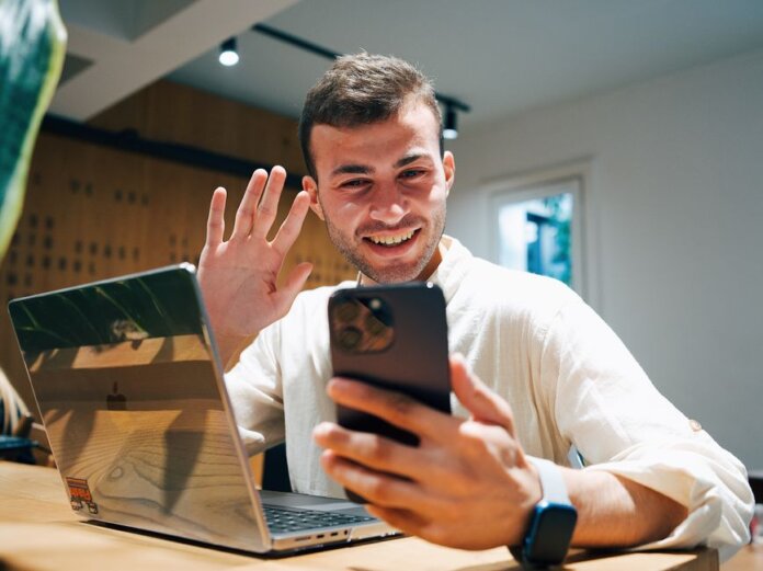 Smiling young man making a video call on his smartphone while sitting with a laptop in a modern office. Smiling young man making a video call on his smartphone while sitting with a laptop in a modern office.