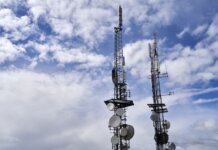 Telecommunication towers with antennas and dishes under a bright cloudy sky.