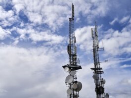 Telecommunication towers with antennas and dishes under a bright cloudy sky.