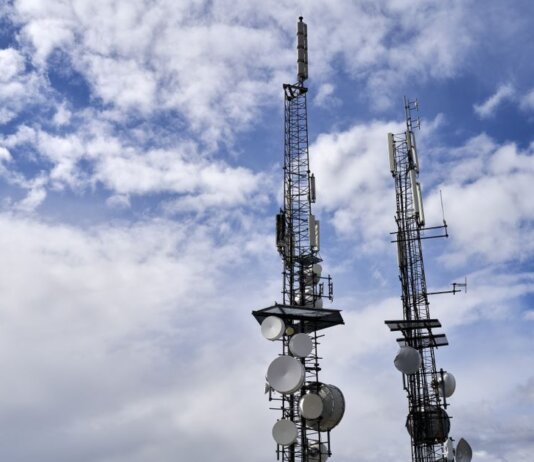 Telecommunication towers with antennas and dishes under a bright cloudy sky.
