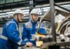 Two industrial workers in protective gear operate machinery in a factory setting.